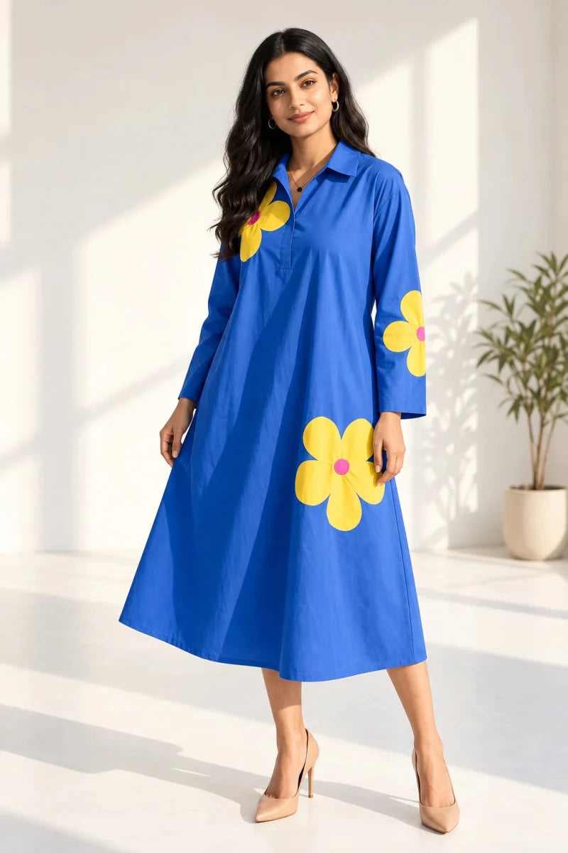 A woman modeling a bright blue midi shirt dress with large yellow flower prints on the front and sleeves, standing in a sunlit room with a potted plant in the background.